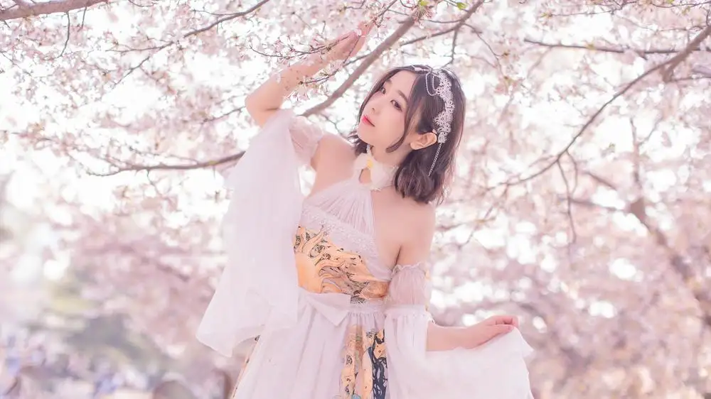 穆零Mu0在樱花树下抬手触碰花枝,白色纱衣与银色发饰,背景为大片粉色花朵