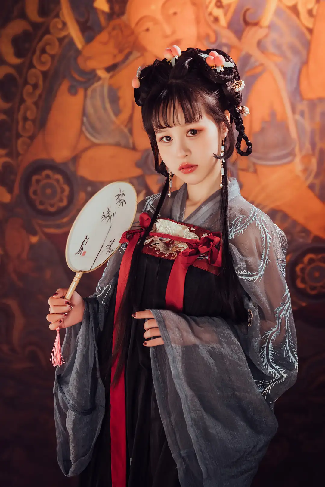 晕崽身穿灰纱大袖衫与红色系带的汉服造型,手持团扇站在敦煌风格壁画前