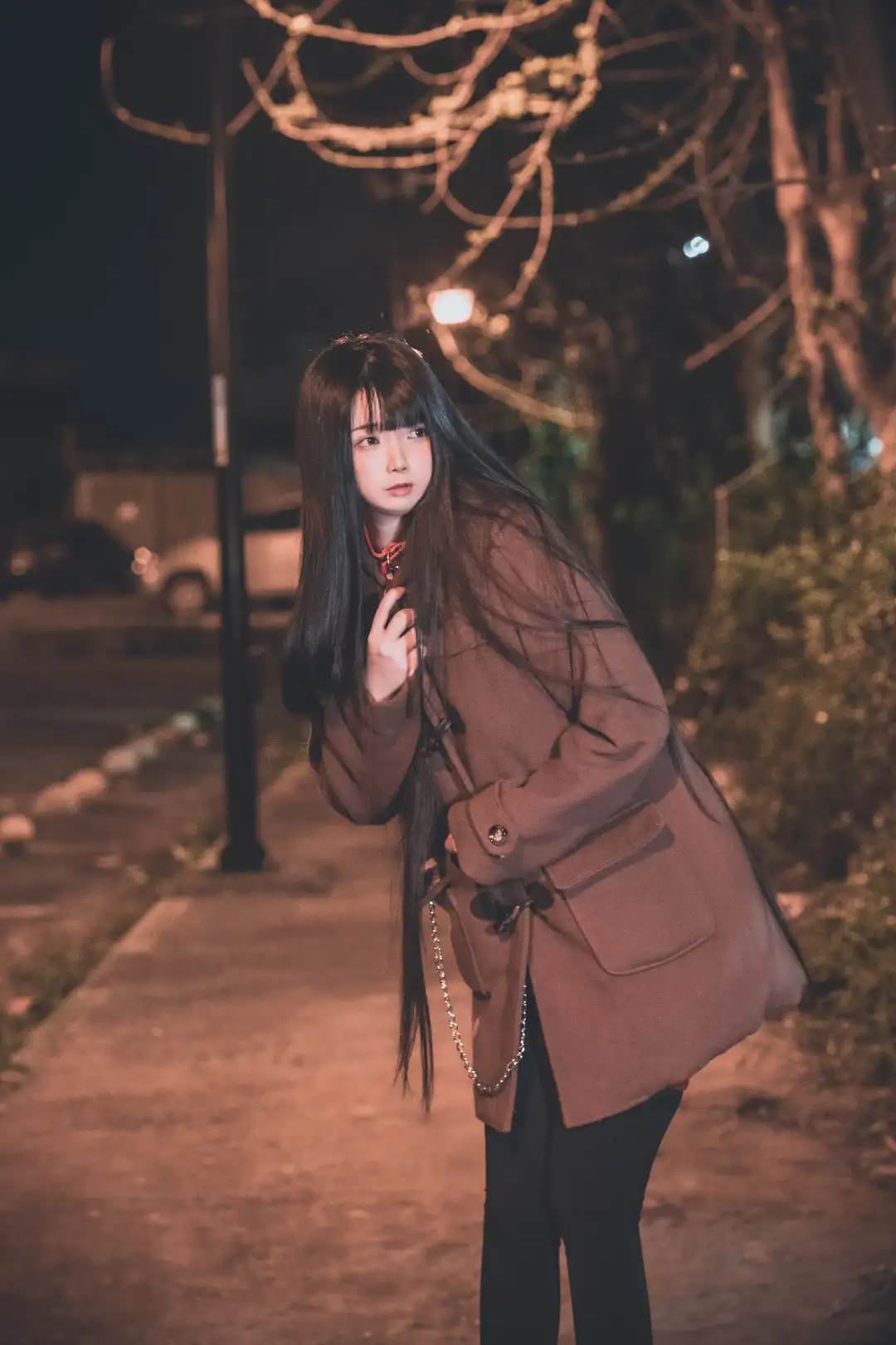 落落Raku夜晚站在街边,穿棕色长外套与黑色长裤,暖色路灯和枝条在背景散景中